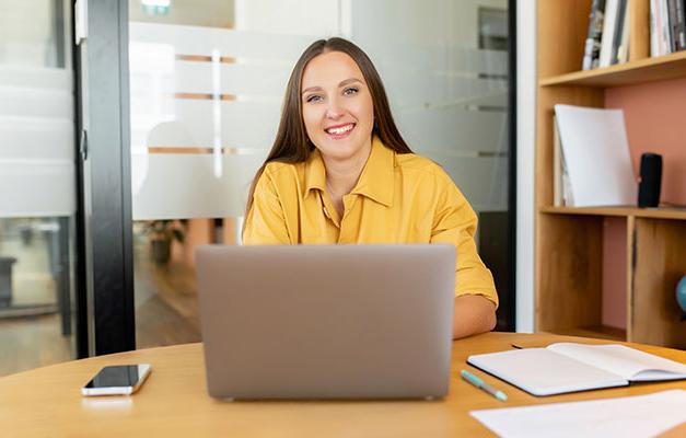 smiling woman in yellow shirt sitting behing a laptop computer in an office